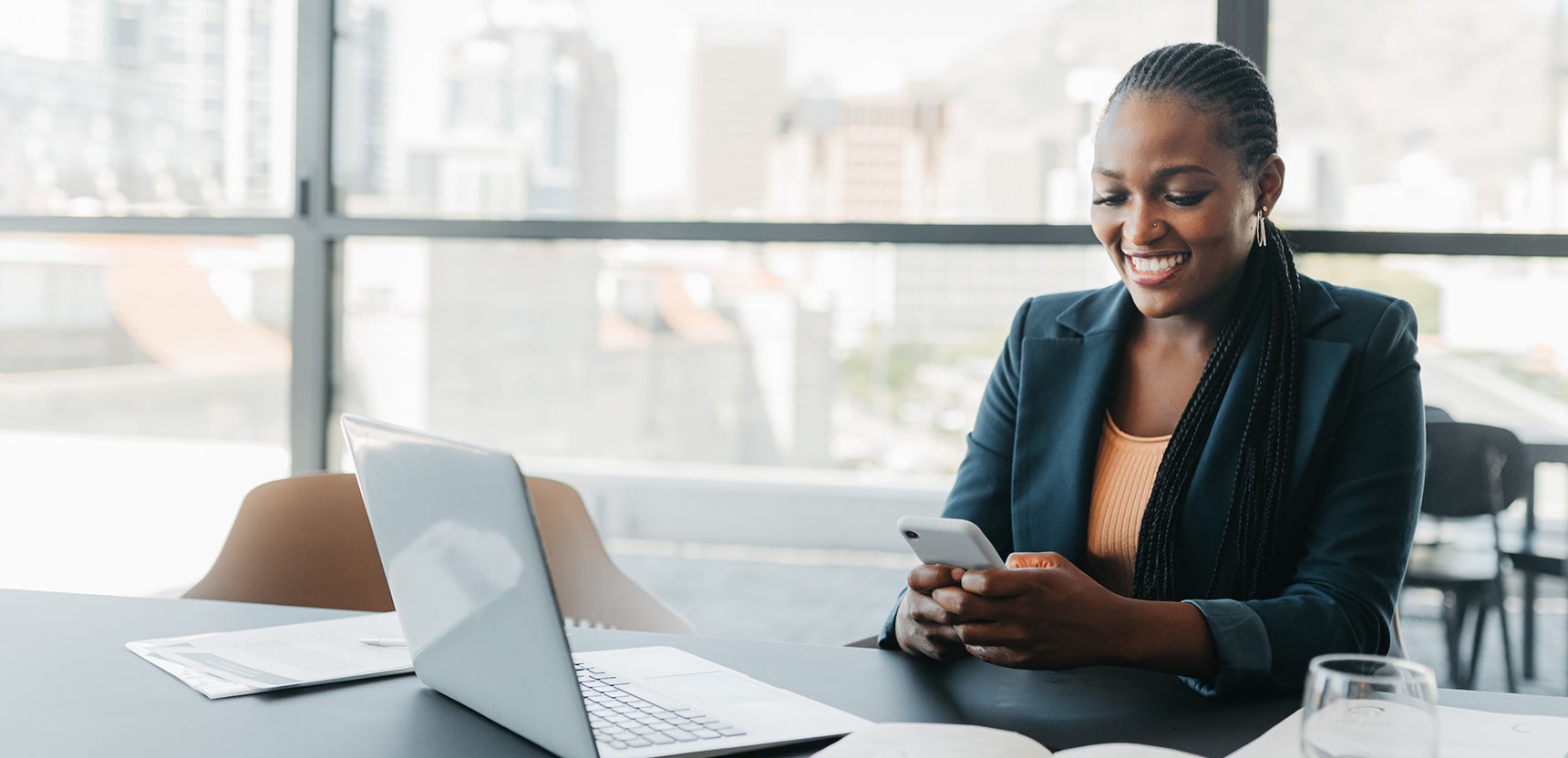 Professional woman in an office with her laptop open checking her smartphone and smiling.