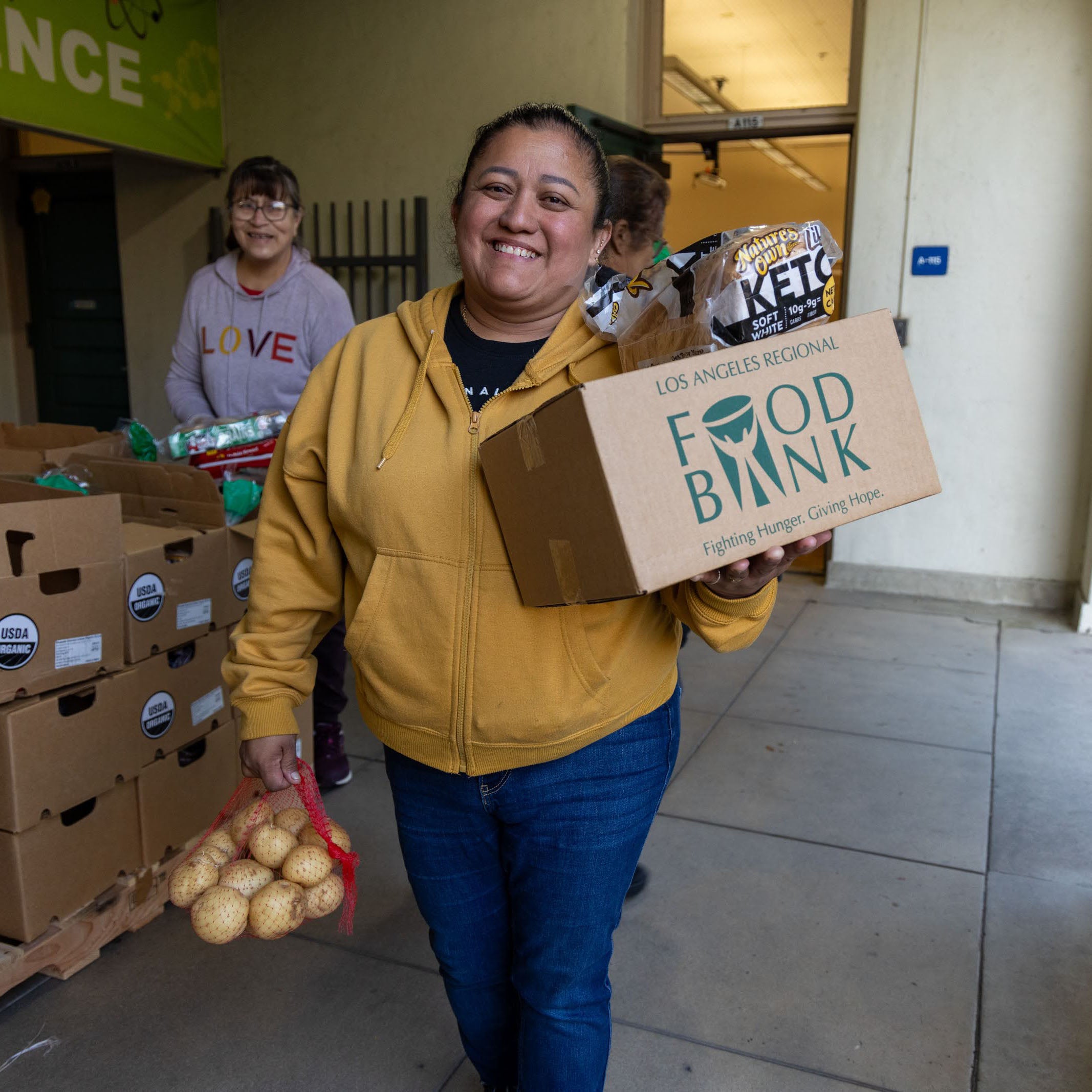 Smiling volunteer in a yellow hoodie carrying a food bank donation box and a bag of potatoes at a distribution site.